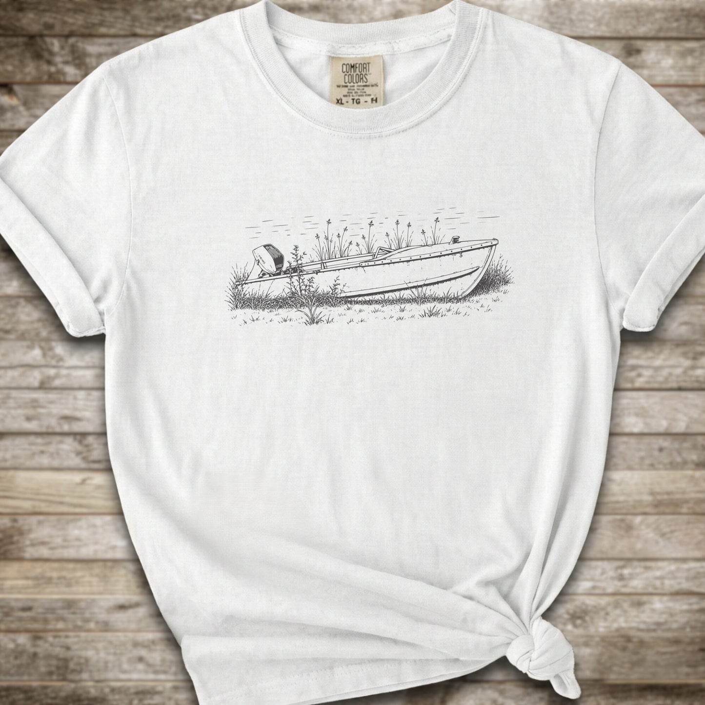 A white short-sleeve crewneck t-shirt with a black line art graphic of a small boat on a marshy shore. The t-shirt is styled with a knot at the bottom hem and is laid flat on a rustic wooden plank background.