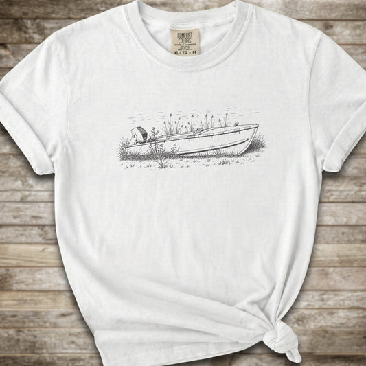 A white short-sleeve crewneck t-shirt with a black line art graphic of a small boat on a marshy shore. The t-shirt is styled with a knot at the bottom hem and is laid flat on a rustic wooden plank background.