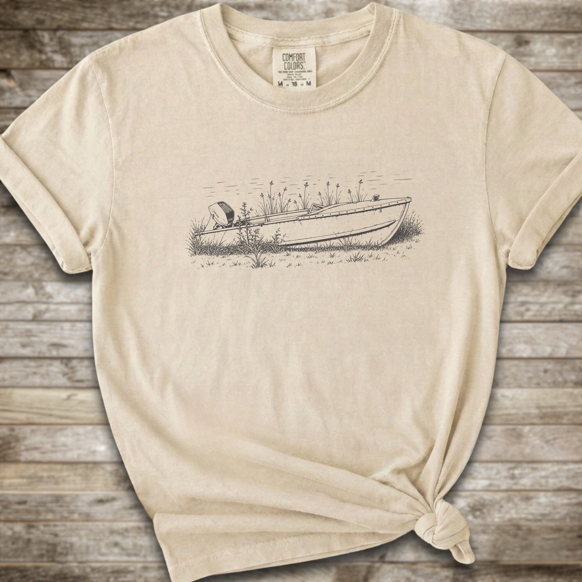 An ivory short-sleeve crewneck t-shirt with a line art graphic of a small boat on a marshy shore. The t-shirt is styled with a knot at the bottom hem and is laid flat on a rustic wooden plank background.