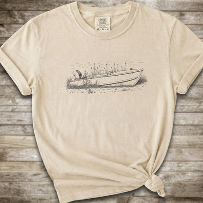 An ivory short-sleeve crewneck t-shirt with a line art graphic of a small boat on a marshy shore. The t-shirt is styled with a knot at the bottom hem and is laid flat on a rustic wooden plank background.