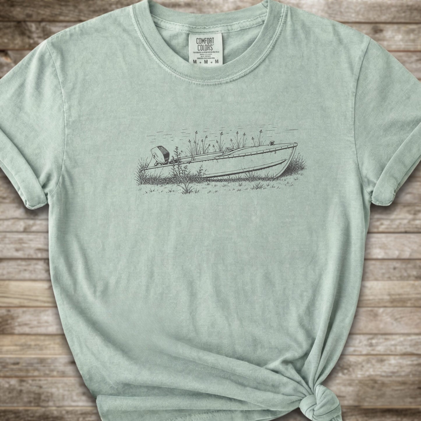 A light green short-sleeve crewneck t-shirt with a line art graphic of a small boat on a marshy shore. The t-shirt is styled with a knot at the bottom hem and is laid flat on a rustic wooden plank background.