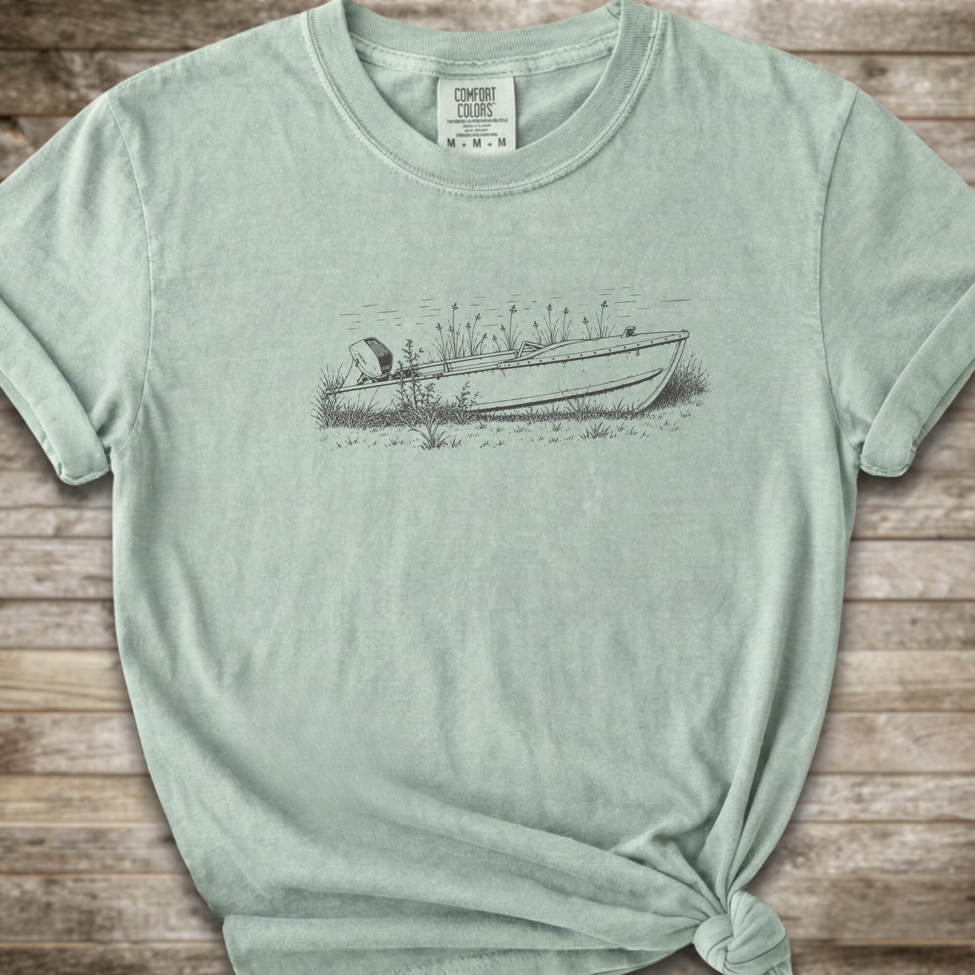 A light green short-sleeve crewneck t-shirt with a line art graphic of a small boat on a marshy shore. The t-shirt is styled with a knot at the bottom hem and is laid flat on a rustic wooden plank background.