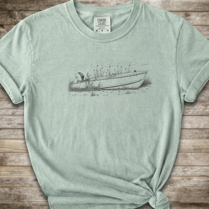 A light green short-sleeve crewneck t-shirt with a line art graphic of a small boat on a marshy shore. The t-shirt is styled with a knot at the bottom hem and is laid flat on a rustic wooden plank background.