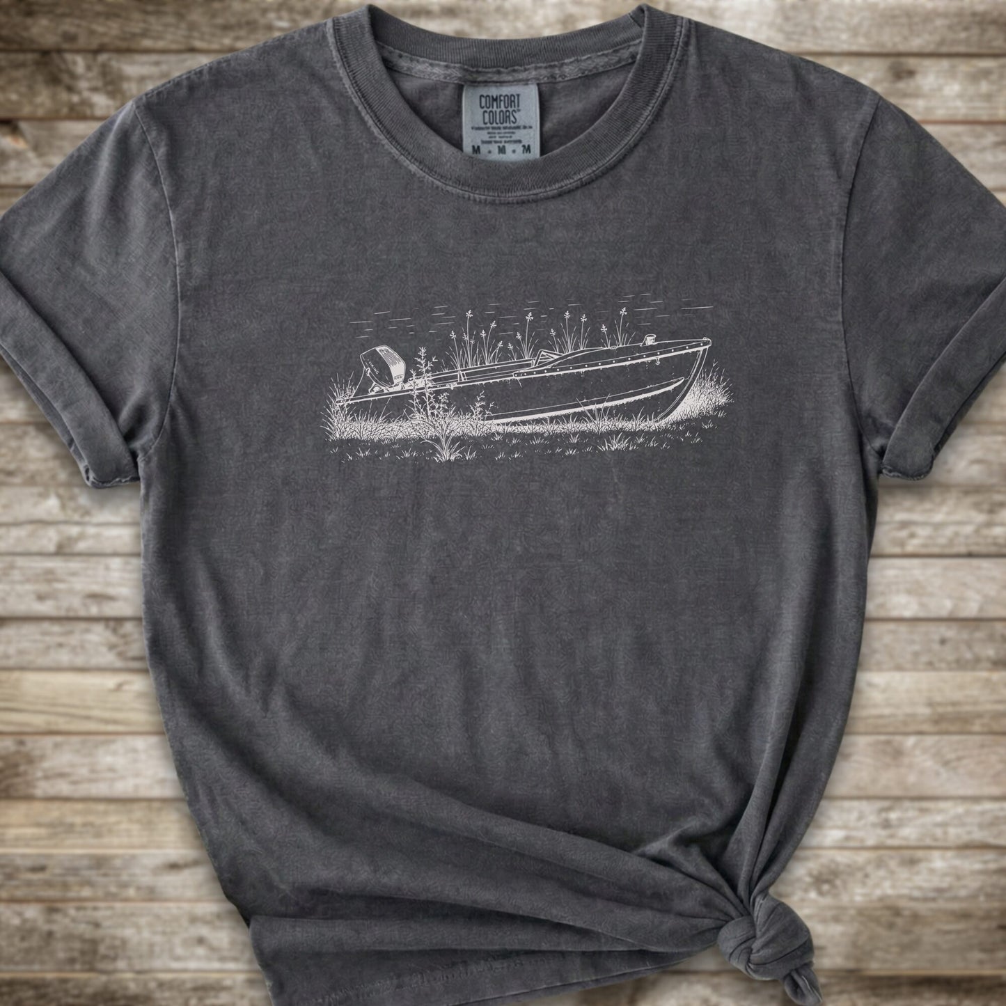 A dark gray t-shirt with a distressed wash, featuring a white line art graphic of a speedboat on a lake. The t-shirt is styled with a knot at the bottom hem and is laid flat on a rustic wooden plank background.