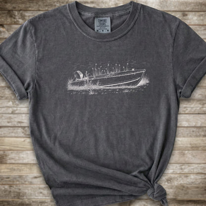 A dark gray t-shirt with a distressed wash, featuring a white line art graphic of a speedboat on a lake. The t-shirt is styled with a knot at the bottom hem and is laid flat on a rustic wooden plank background.
