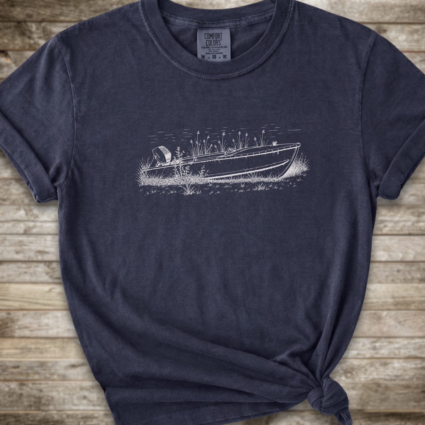 A navy short-sleeve crewneck t-shirt with a line art graphic of a small boat on a marshy shore. The t-shirt is styled with a knot at the bottom hem and is laid flat on a rustic wooden plank background.