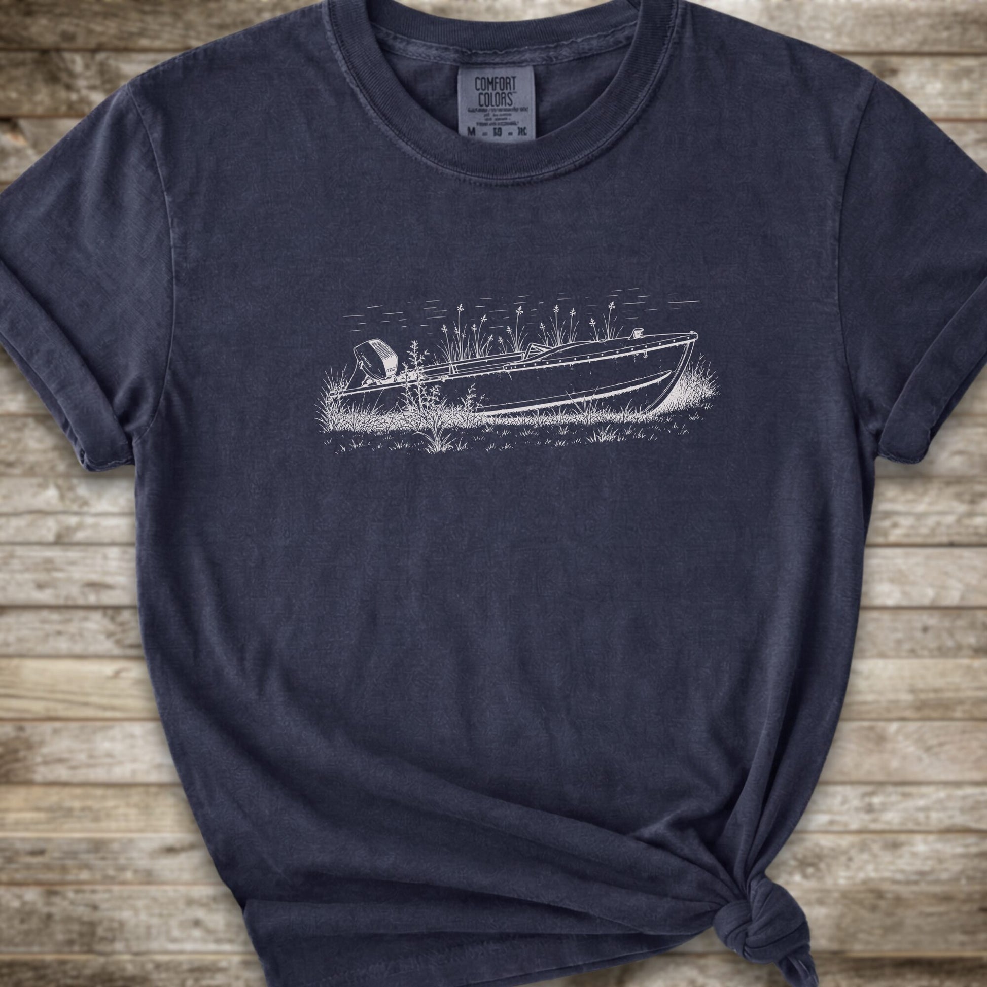 A navy short-sleeve crewneck t-shirt with a line art graphic of a small boat on a marshy shore. The t-shirt is styled with a knot at the bottom hem and is laid flat on a rustic wooden plank background.