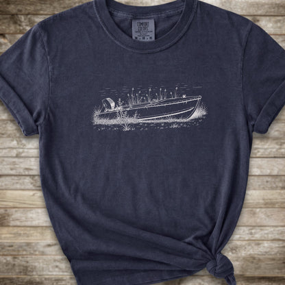 A navy short-sleeve crewneck t-shirt with a line art graphic of a small boat on a marshy shore. The t-shirt is styled with a knot at the bottom hem and is laid flat on a rustic wooden plank background.