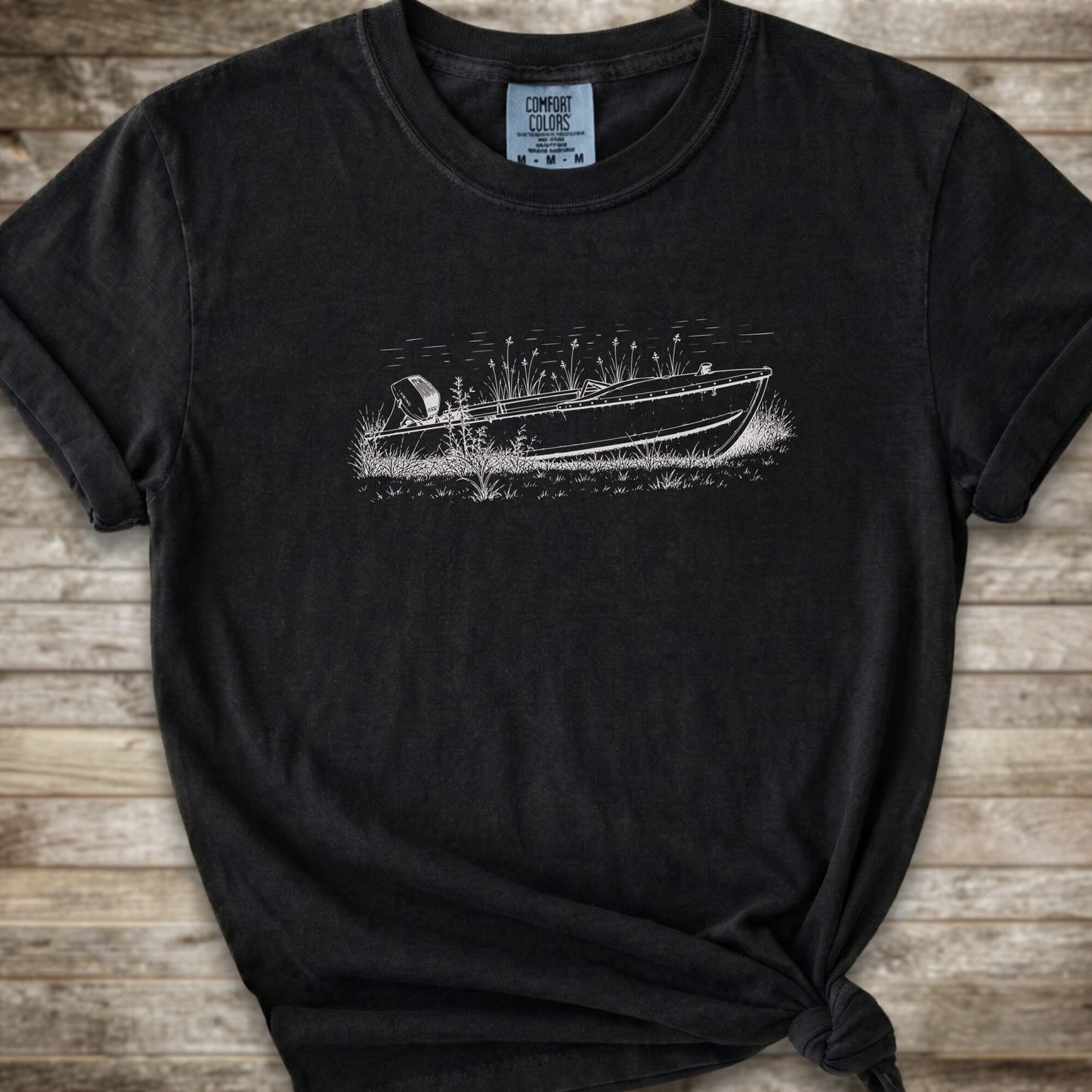 A black short-sleeve crewneck t-shirt with a white line art graphic of a small boat on a marshy shore. The t-shirt is styled with a knot at the bottom hem and is laid flat on a rustic wooden plank background.