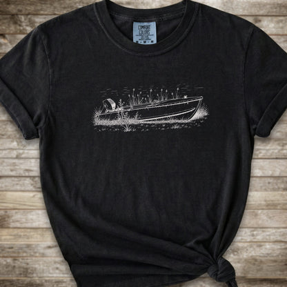 A black short-sleeve crewneck t-shirt with a white line art graphic of a small boat on a marshy shore. The t-shirt is styled with a knot at the bottom hem and is laid flat on a rustic wooden plank background.