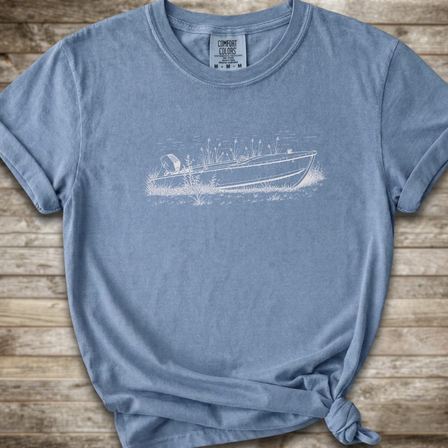 A blue short-sleeve crewneck t-shirt with a white  line art graphic of a small boat on a marshy shore. The t-shirt is styled with a knot at the bottom hem and is laid flat on a rustic wooden plank background.