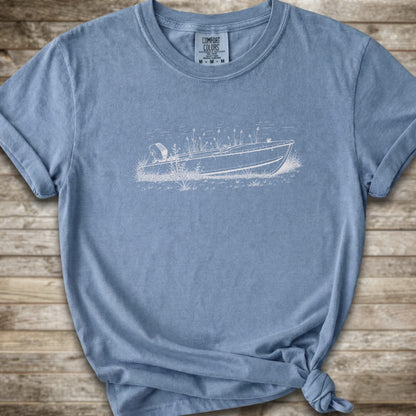 A blue short-sleeve crewneck t-shirt with a white  line art graphic of a small boat on a marshy shore. The t-shirt is styled with a knot at the bottom hem and is laid flat on a rustic wooden plank background.