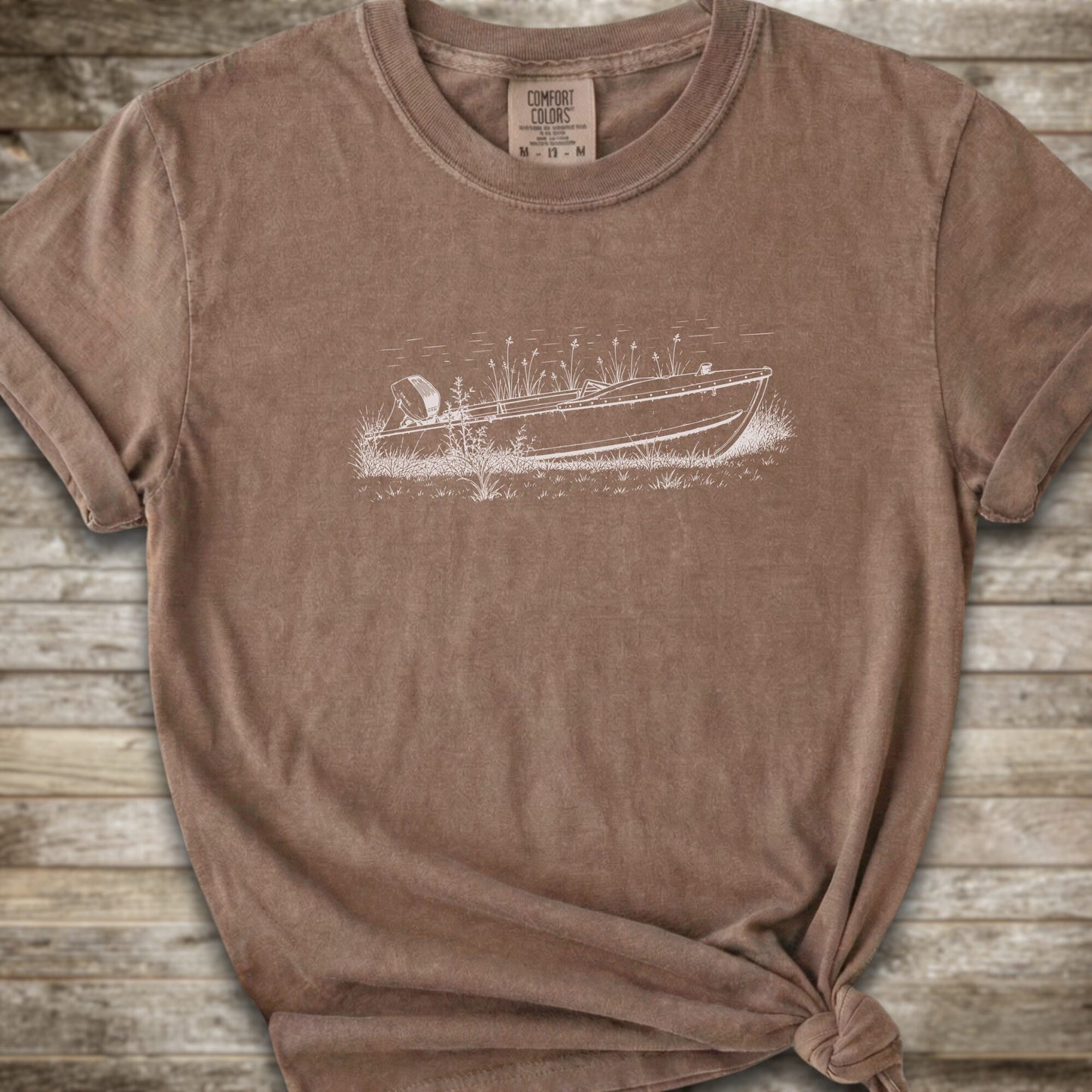 A brown short-sleeve crewneck t-shirt with white line art graphic of a small boat on a marshy shore. The t-shirt is styled with a knot at the bottom hem and is laid flat on a rustic wooden plank background.