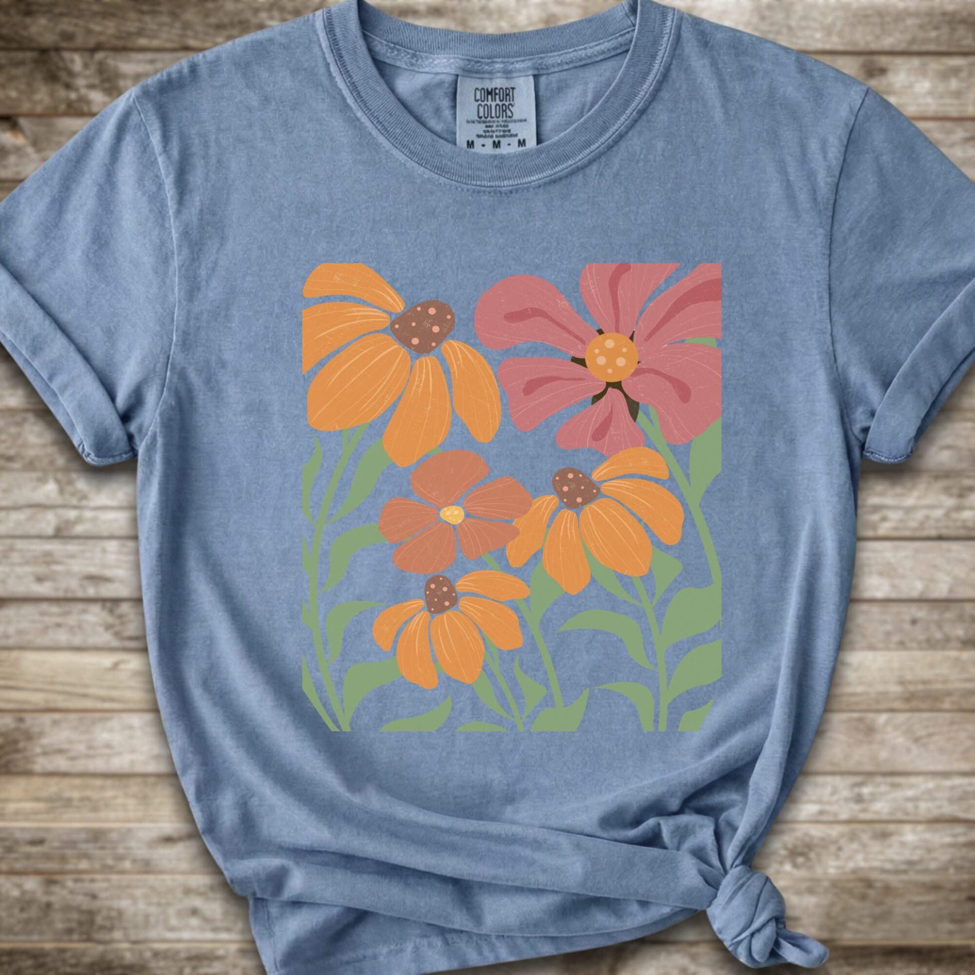 A light Blue short-sleeve t-shirt with a graphic print of stylized orange, pink, and brown flowers with green leaves, styled on a rustic wooden background with a knot at the bottom hem.