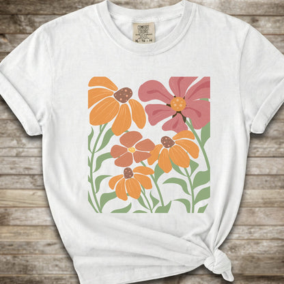 A white short-sleeve t-shirt with a graphic print of stylized orange, pink, and brown flowers with green leaves, styled on a rustic wooden background with a knot at the bottom hem.