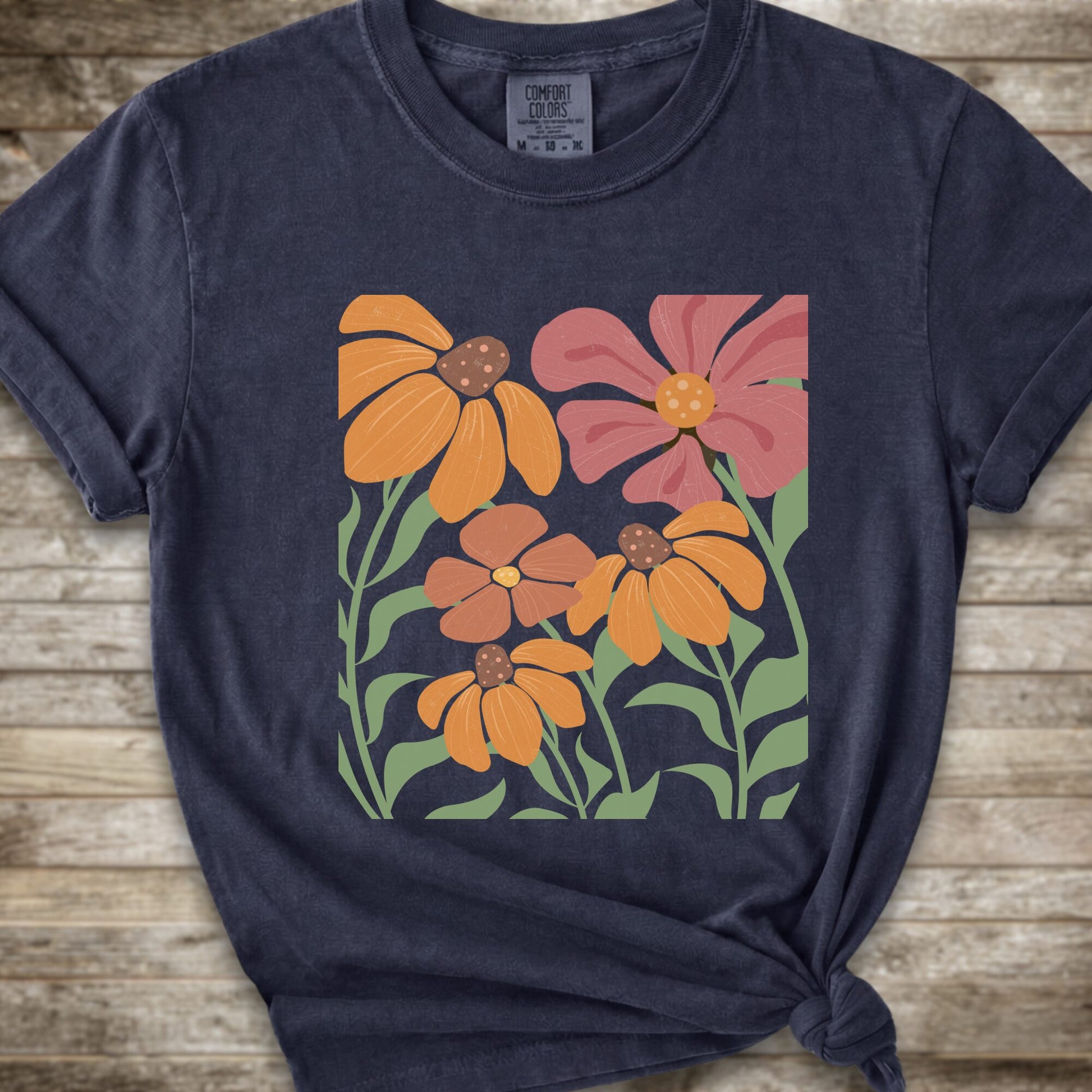 A Navy Blue short-sleeve t-shirt with a graphic print of stylized orange, pink, and brown flowers with green leaves, styled on a rustic wooden background with a knot at the bottom hem.