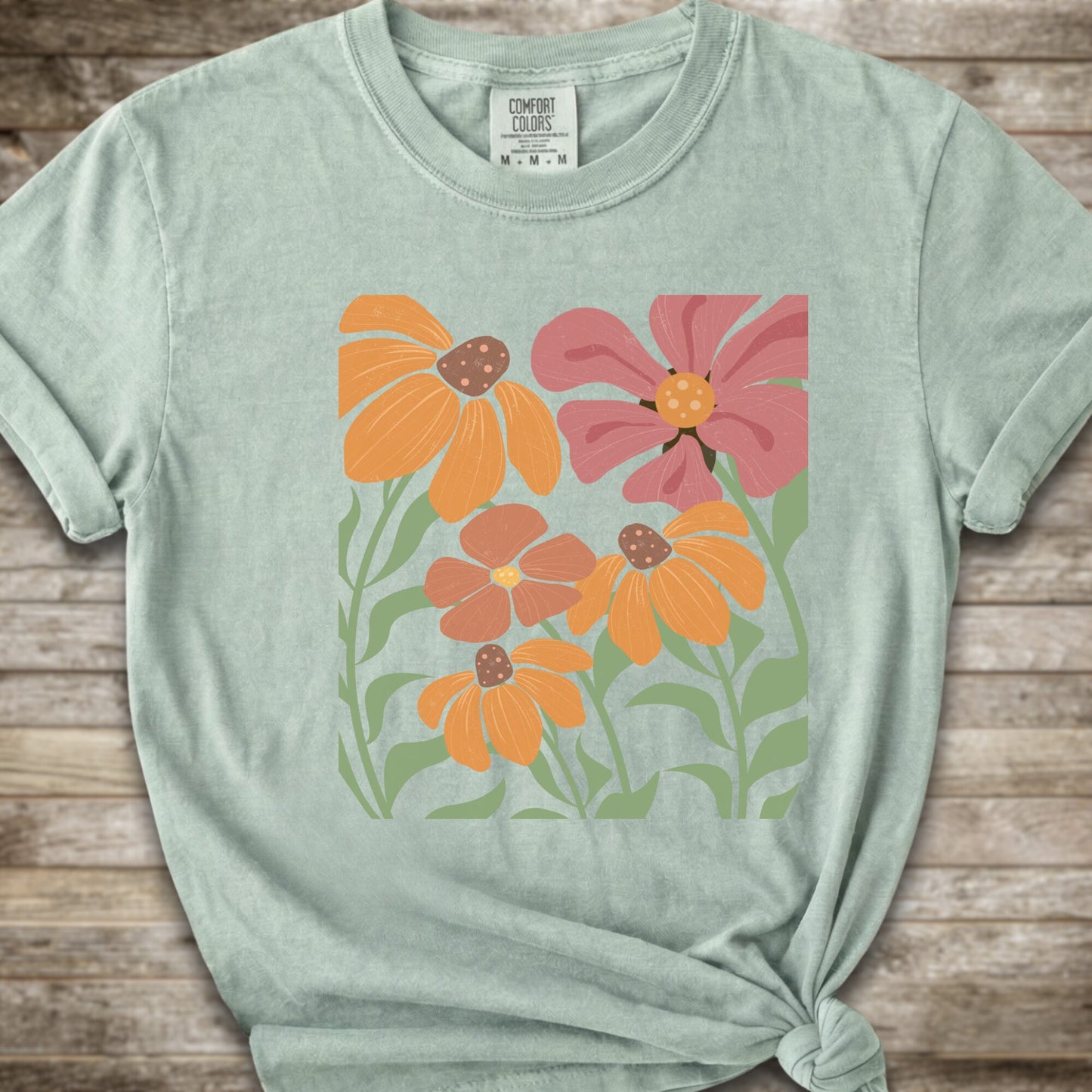 A Litght Green short-sleeve t-shirt with a graphic print of stylized orange, pink, and brown flowers with green leaves, styled on a rustic wooden background with a knot at the bottom hem.