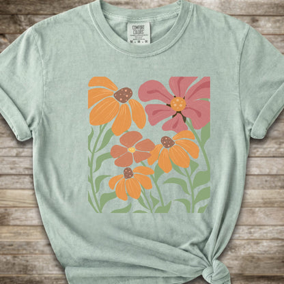 A Litght Green short-sleeve t-shirt with a graphic print of stylized orange, pink, and brown flowers with green leaves, styled on a rustic wooden background with a knot at the bottom hem.