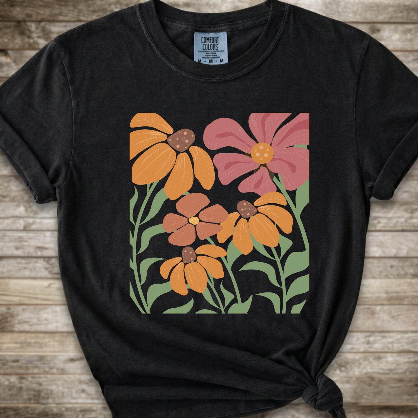 A Black short-sleeve t-shirt with a graphic print of stylized orange, pink, and brown flowers with green leaves, styled on a rustic wooden background with a knot at the bottom hem.