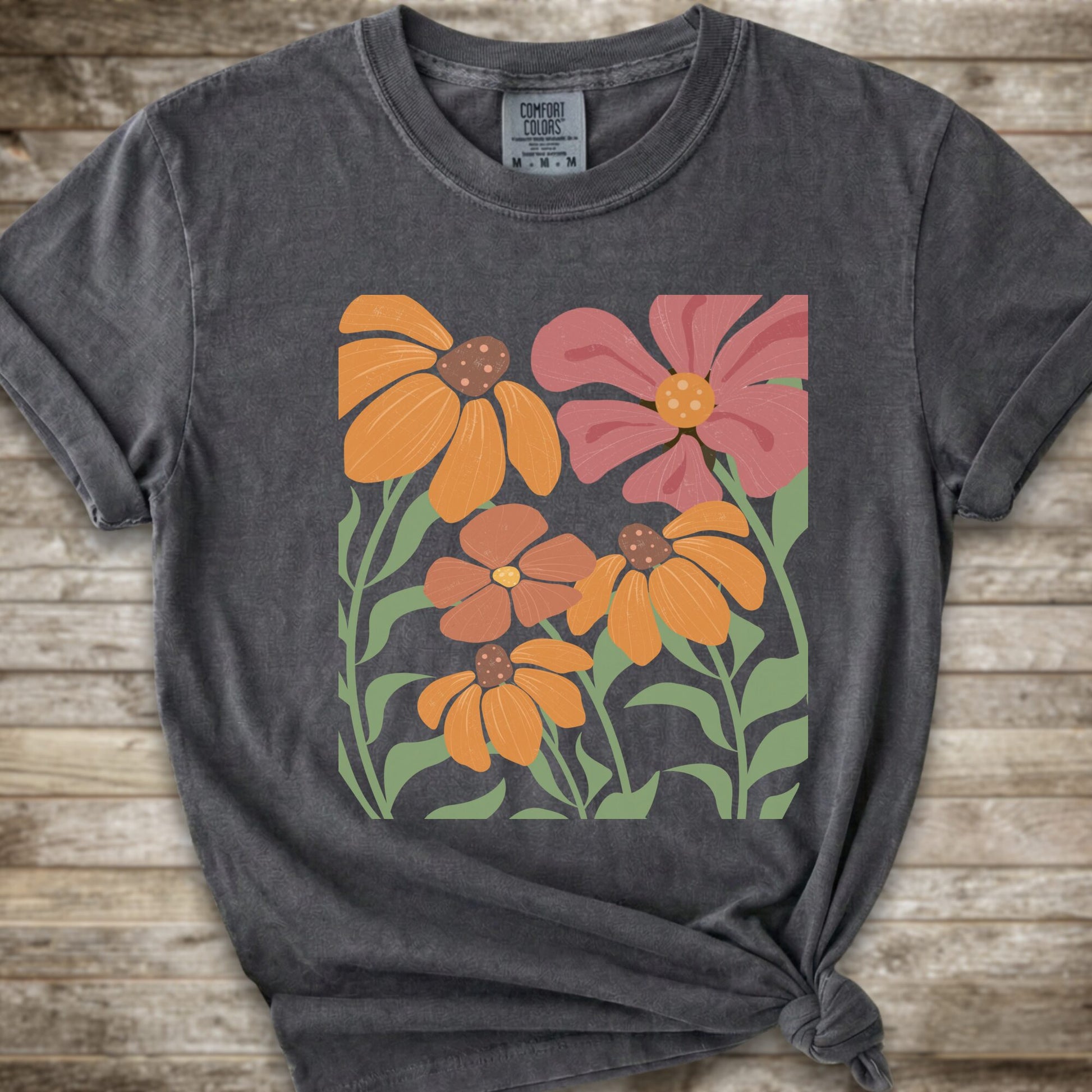 A Dark Grey short-sleeve t-shirt with a graphic print of stylized orange, pink, and brown flowers with green leaves, styled on a rustic wooden background with a knot at the bottom hem.