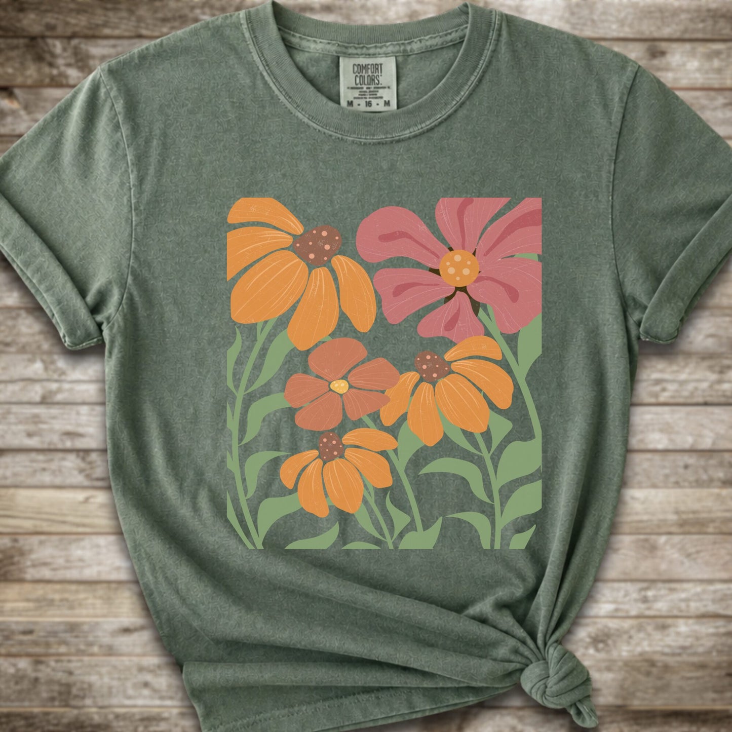 A Moss Green short-sleeve t-shirt with a graphic print of stylized orange, pink, and brown flowers with green leaves, styled on a rustic wooden background with a knot at the bottom hem.