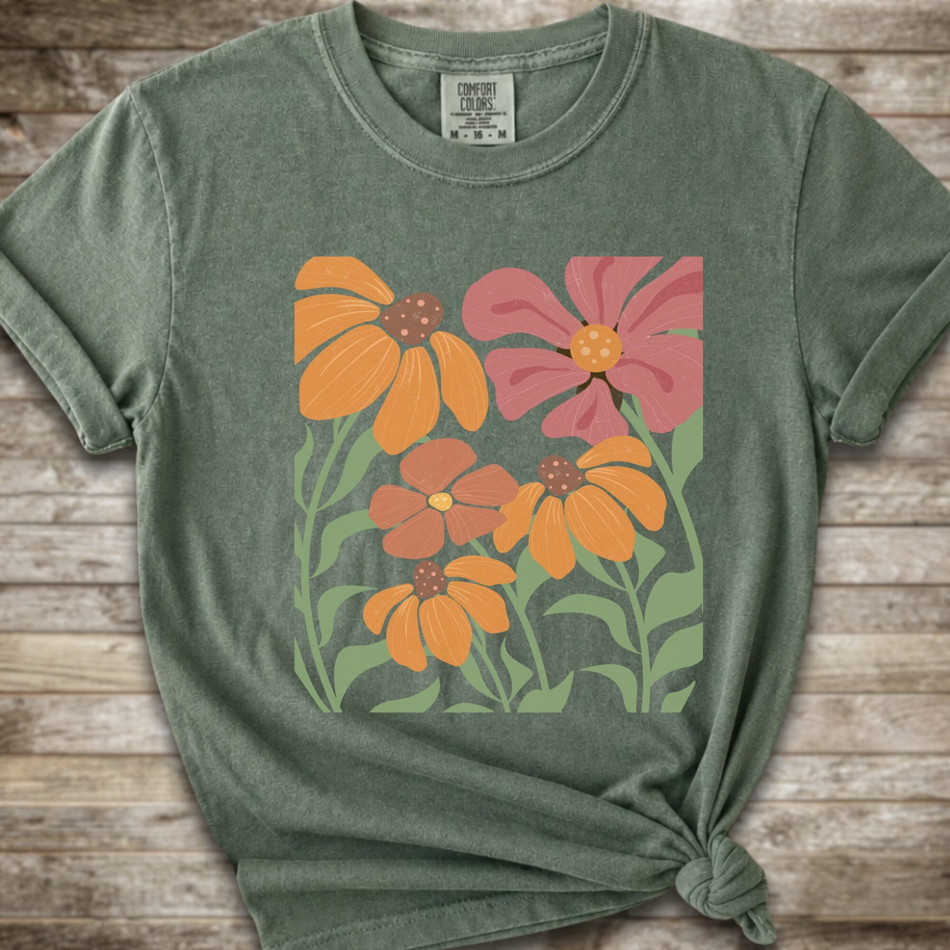 A Moss Green short-sleeve t-shirt with a graphic print of stylized orange, pink, and brown flowers with green leaves, styled on a rustic wooden background with a knot at the bottom hem.
