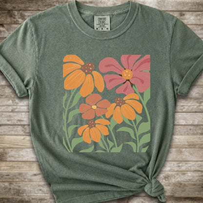 A Moss Green short-sleeve t-shirt with a graphic print of stylized orange, pink, and brown flowers with green leaves, styled on a rustic wooden background with a knot at the bottom hem.