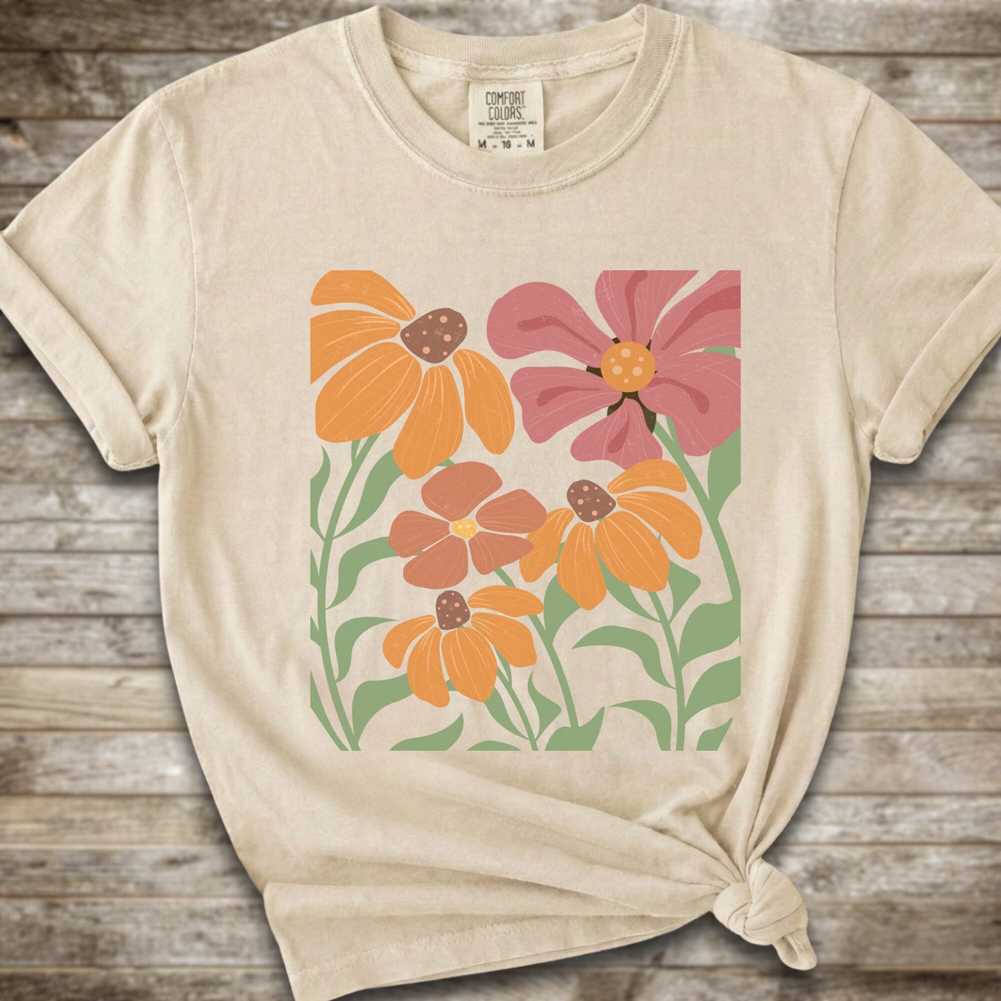 An Ivory short-sleeve t-shirt with a graphic print of stylized orange, pink, and brown flowers with green leaves, styled on a rustic wooden background with a knot at the bottom hem.
