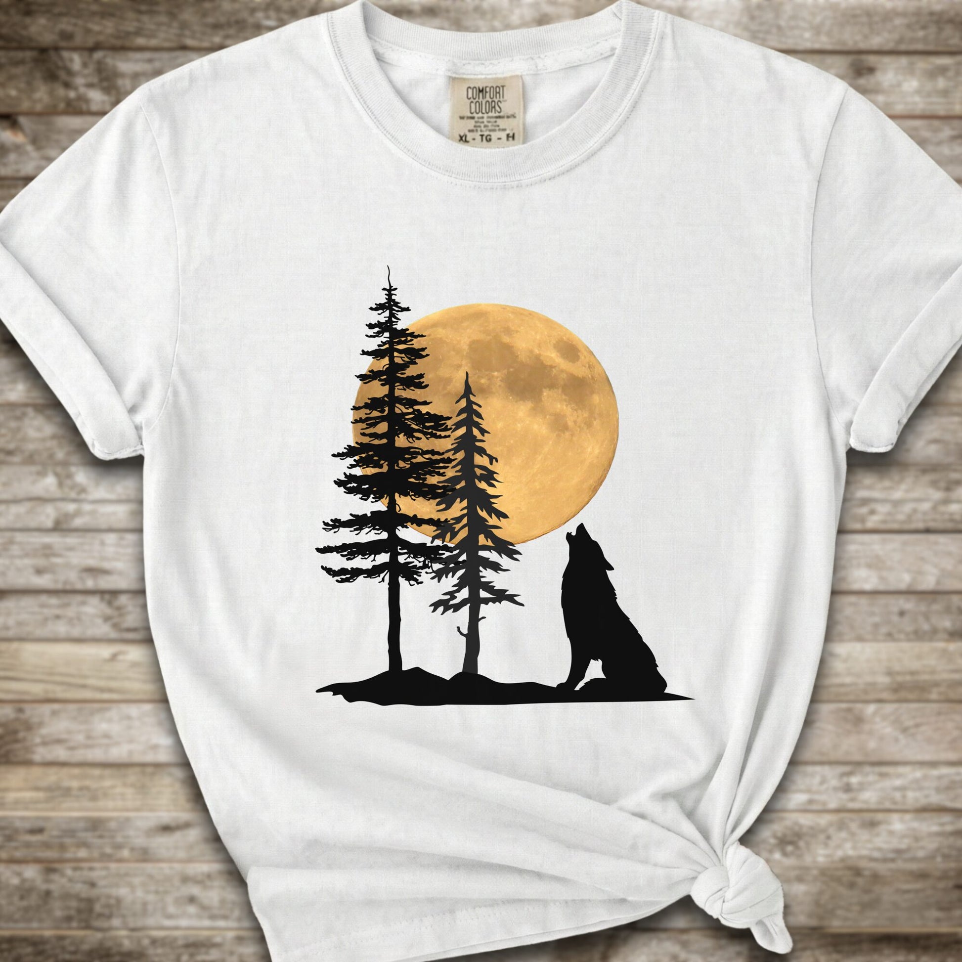 A white short-sleeve t-shirt with a graphic of a black wolf silhouette howling in front of two pine trees and a large yellow full moon, displayed on a rustic wooden background.
