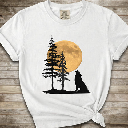 A white short-sleeve t-shirt with a graphic of a black wolf silhouette howling in front of two pine trees and a large yellow full moon, displayed on a rustic wooden background.