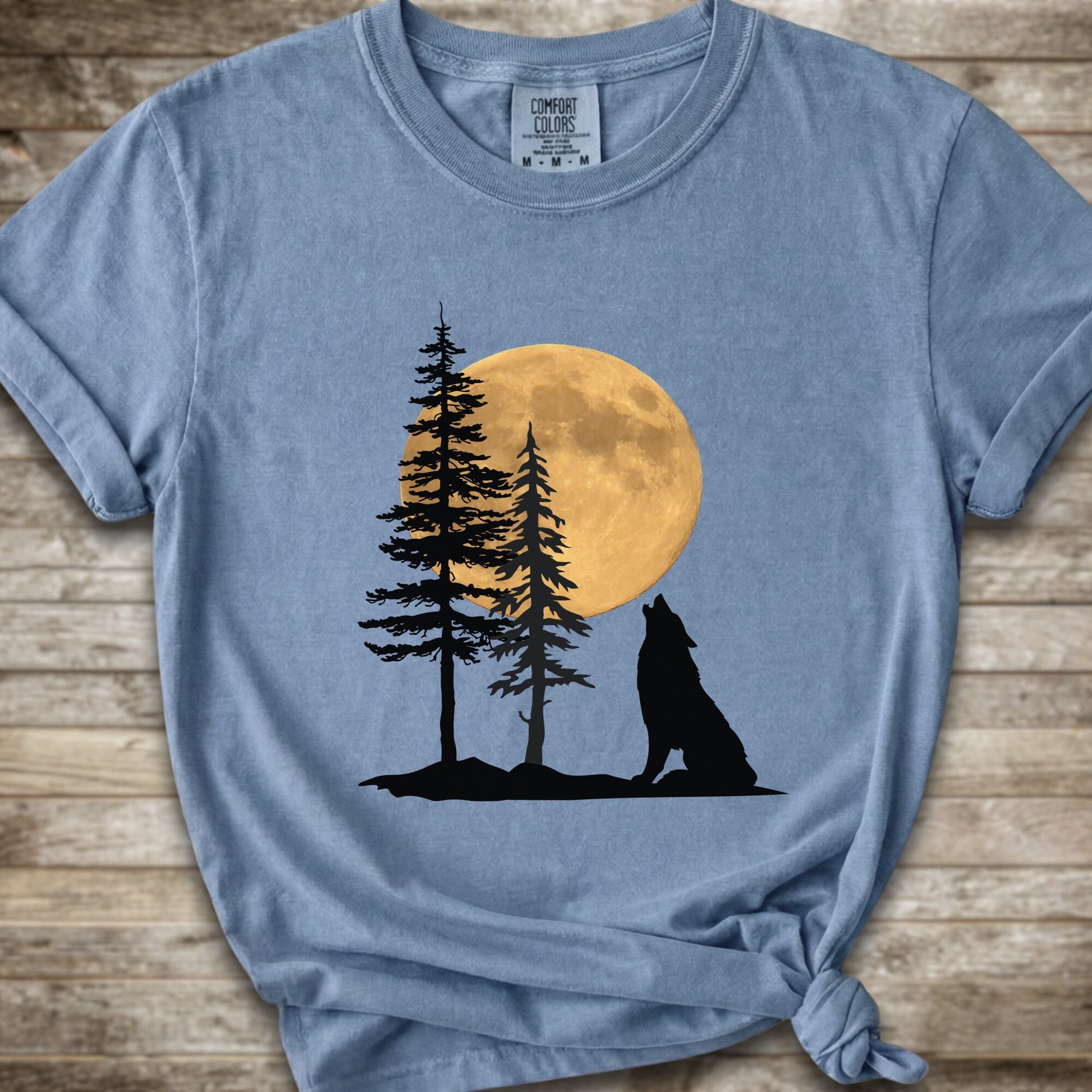 A light blue short-sleeve t-shirt with a graphic of a black wolf silhouette howling in front of two pine trees and a large yellow full moon, displayed on a rustic wooden background.