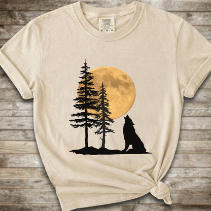 An Ivory short-sleeve t-shirt with a graphic of a black wolf silhouette howling in front of two pine trees and a large yellow full moon, displayed on a rustic wooden background.