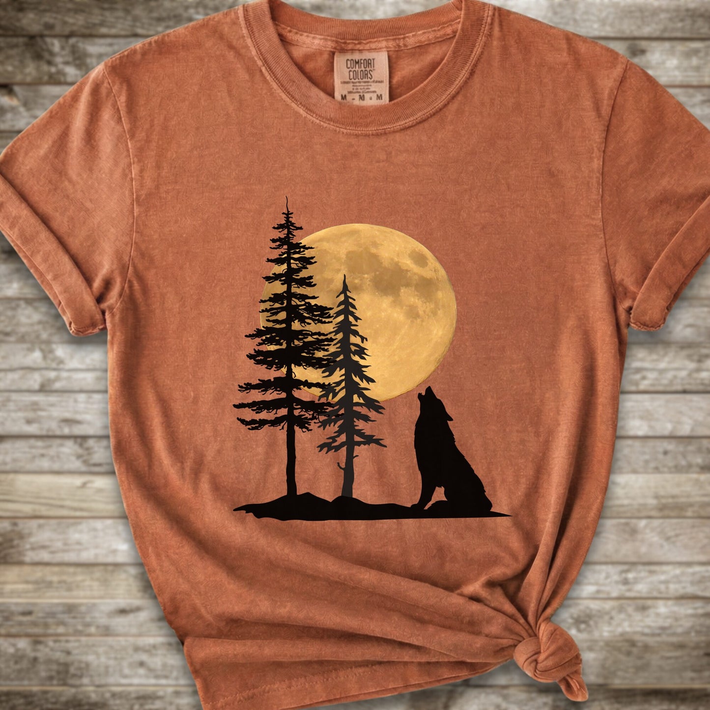 A Burnt Orange short-sleeve t-shirt with a graphic of a black wolf silhouette howling in front of two pine trees and a large yellow full moon, displayed on a rustic wooden background.