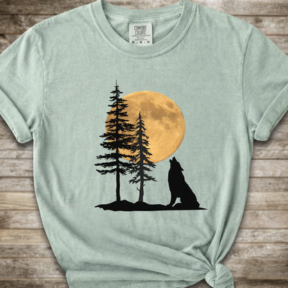 A light green short-sleeve t-shirt with a graphic of a black wolf silhouette howling in front of two pine trees and a large yellow full moon, displayed on a rustic wooden background.