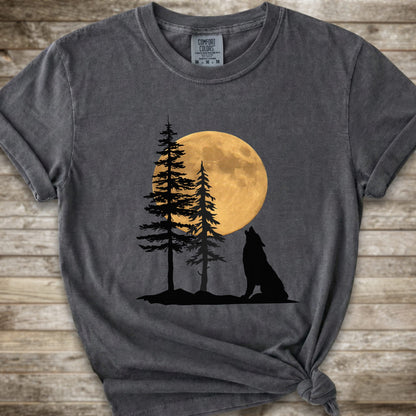 A dark grey short-sleeve t-shirt with a graphic of a black wolf silhouette howling in front of two pine trees and a large yellow full moon, displayed on a rustic wooden background.