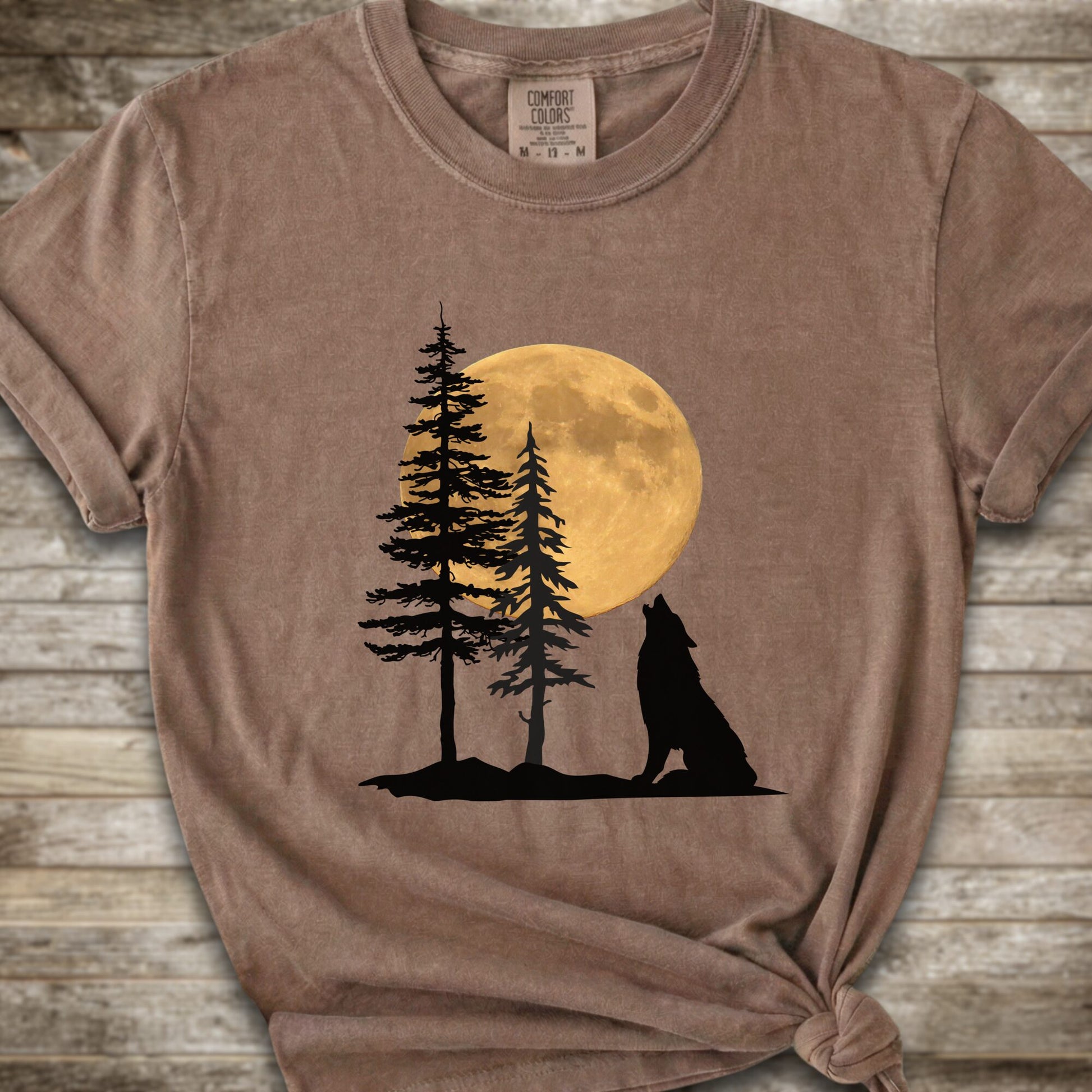 A brown short-sleeve t-shirt with a graphic of a black wolf silhouette howling in front of two pine trees and a large yellow full moon, displayed on a rustic wooden background.
