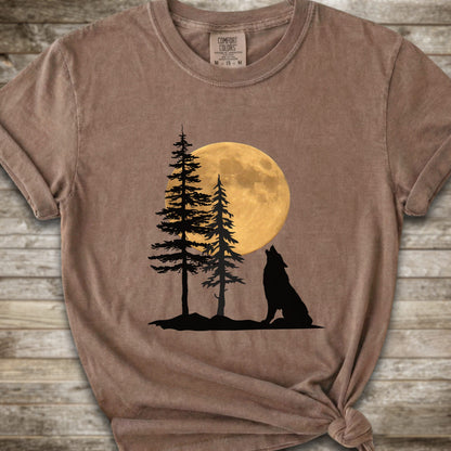 A brown short-sleeve t-shirt with a graphic of a black wolf silhouette howling in front of two pine trees and a large yellow full moon, displayed on a rustic wooden background.