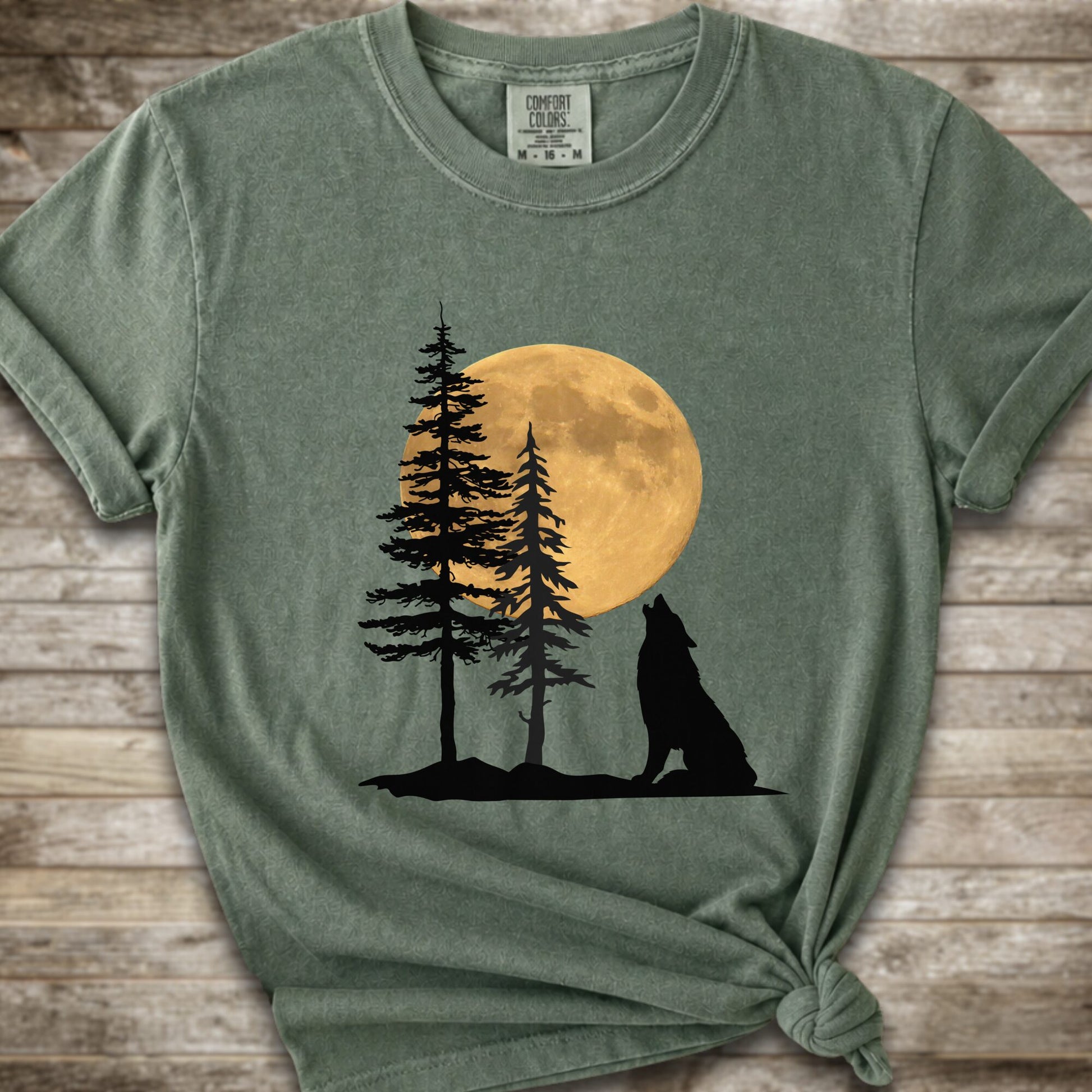 A moss green short-sleeve t-shirt with a graphic of a black wolf silhouette howling in front of two pine trees and a large yellow full moon, displayed on a rustic wooden background.