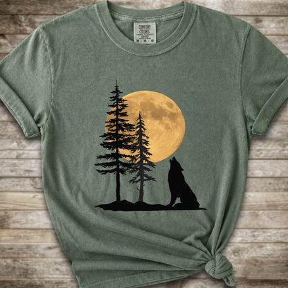 A moss green short-sleeve t-shirt with a graphic of a black wolf silhouette howling in front of two pine trees and a large yellow full moon, displayed on a rustic wooden background.
