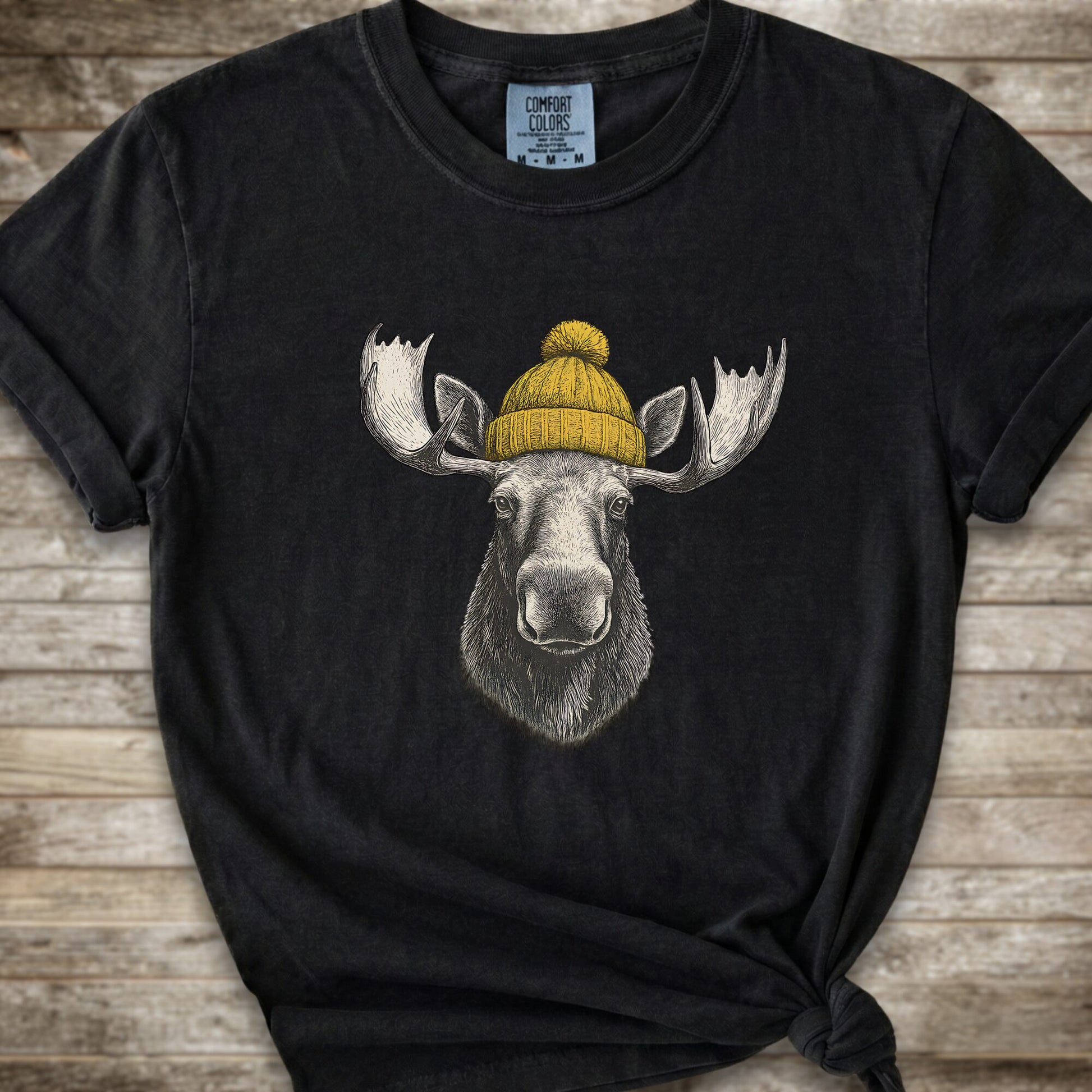 A black short-sleeve t-shirt with a graphic of a moose wearing a yellow knit beanie, laid flat on a rustic wooden background.