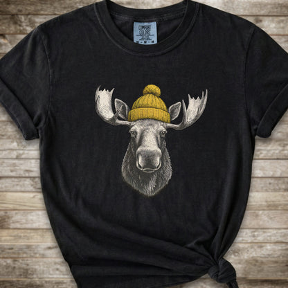 A black short-sleeve t-shirt with a graphic of a moose wearing a yellow knit beanie, laid flat on a rustic wooden background.