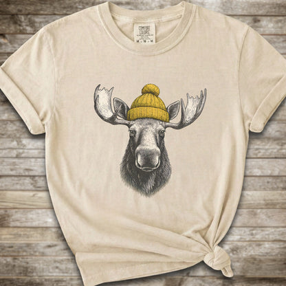 An ivory short-sleeve t-shirt with a graphic of a moose wearing a yellow knit beanie, displayed on a rustic wooden background.