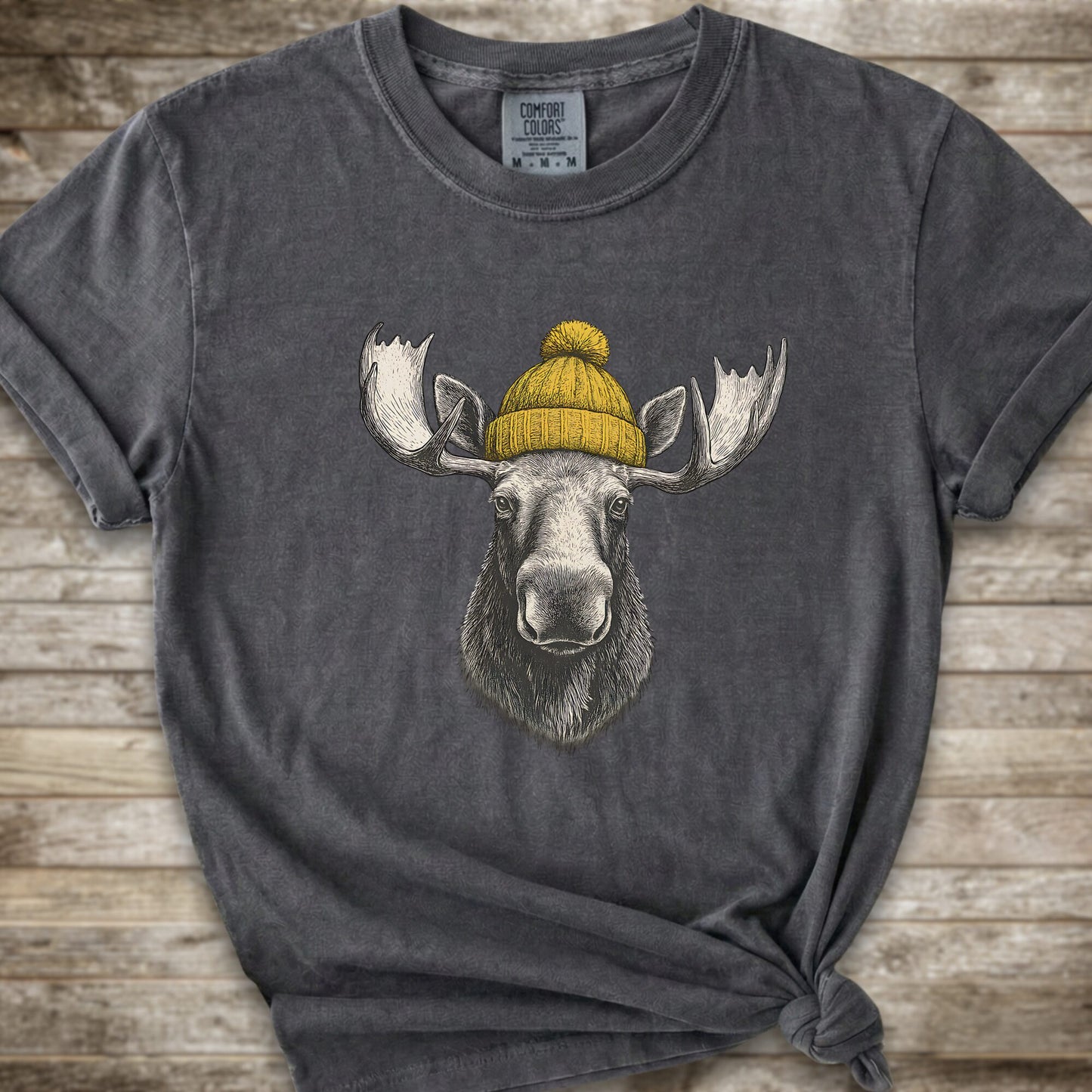 A dark grey short-sleeve t-shirt with a graphic of a moose wearing a yellow knit beanie, displayed on a rustic wooden background.