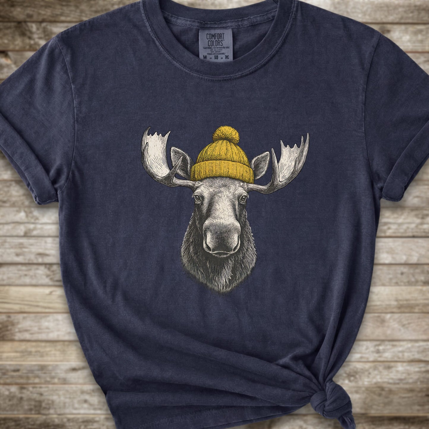 A navy blue short-sleeve t-shirt with a graphic of a moose wearing a yellow knit beanie, displayed on a rustic wooden background.