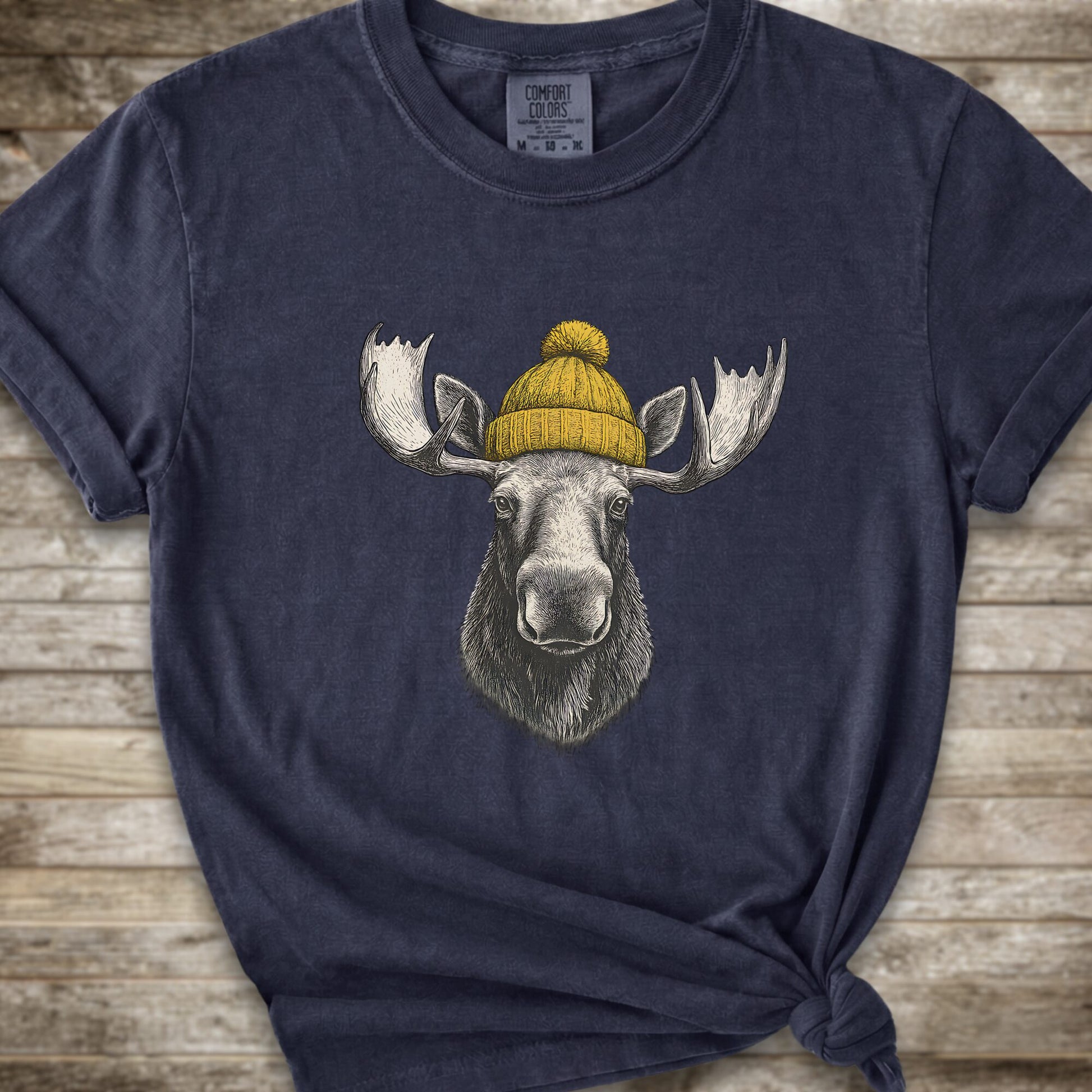 A navy blue short-sleeve t-shirt with a graphic of a moose wearing a yellow knit beanie, displayed on a rustic wooden background.