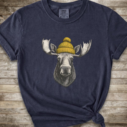 A navy blue short-sleeve t-shirt with a graphic of a moose wearing a yellow knit beanie, displayed on a rustic wooden background.