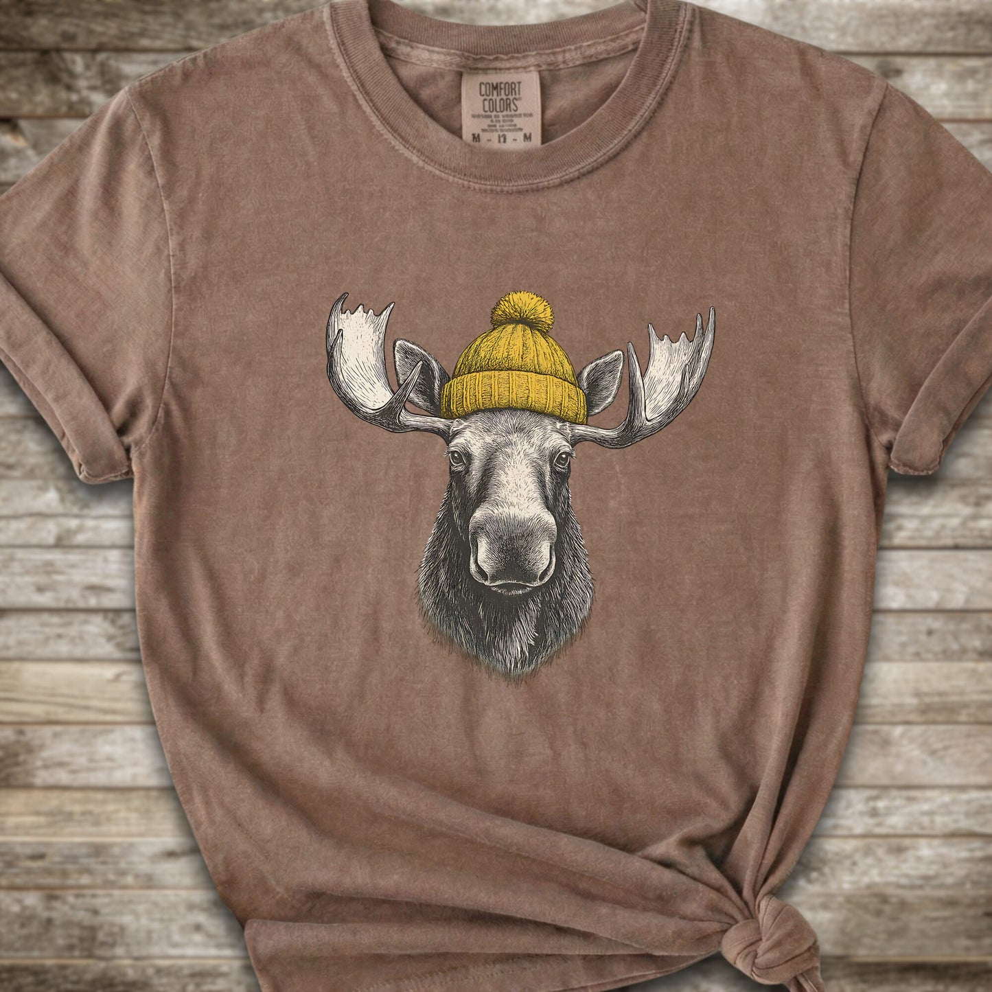 A brown short-sleeve t-shirt with a graphic of a moose wearing a yellow knit beanie, displayed on a rustic wooden background.
