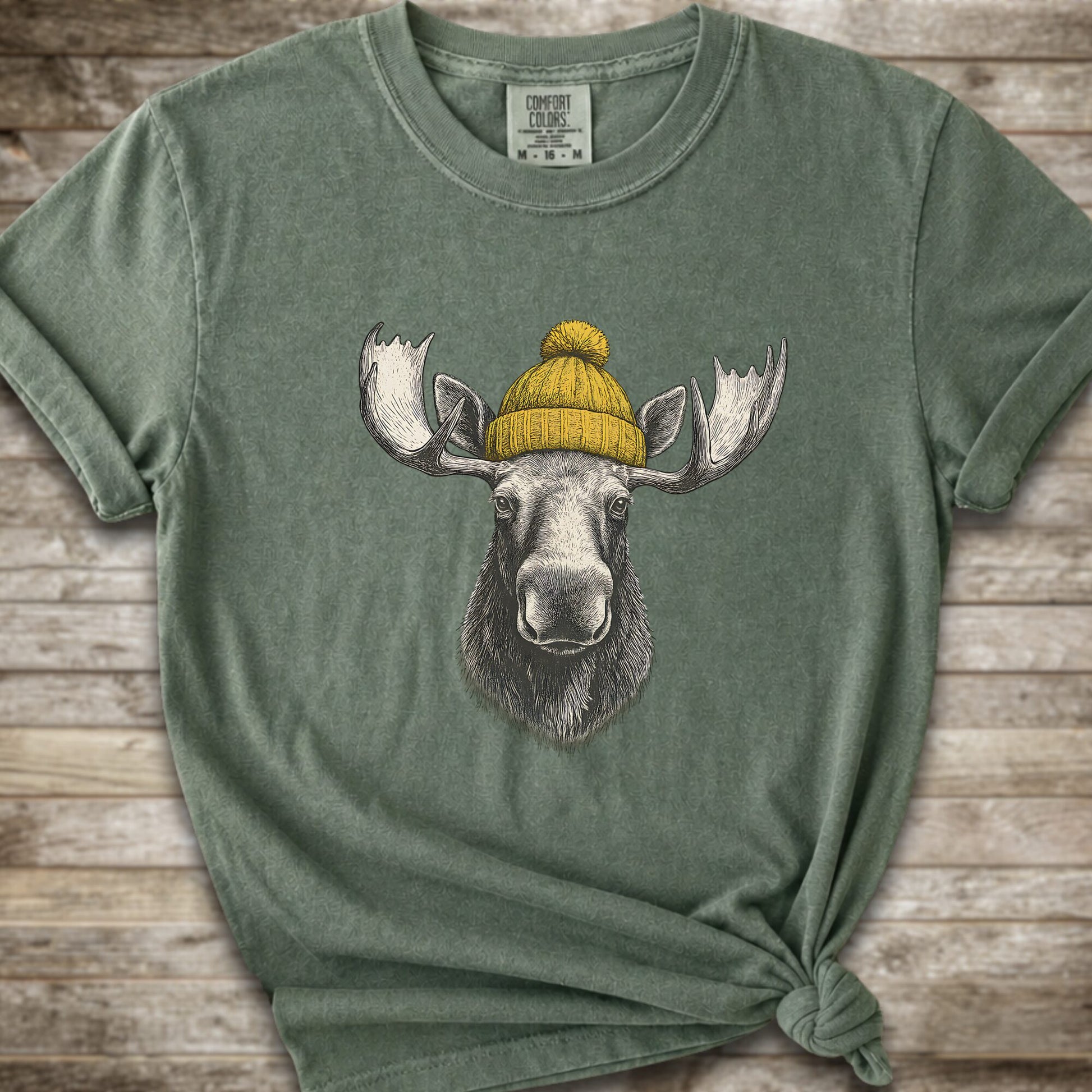 A moss green short-sleeve t-shirt with a graphic of a moose wearing a yellow knit beanie, displayed on a rustic wooden background.