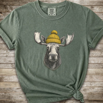 A moss green short-sleeve t-shirt with a graphic of a moose wearing a yellow knit beanie, displayed on a rustic wooden background.