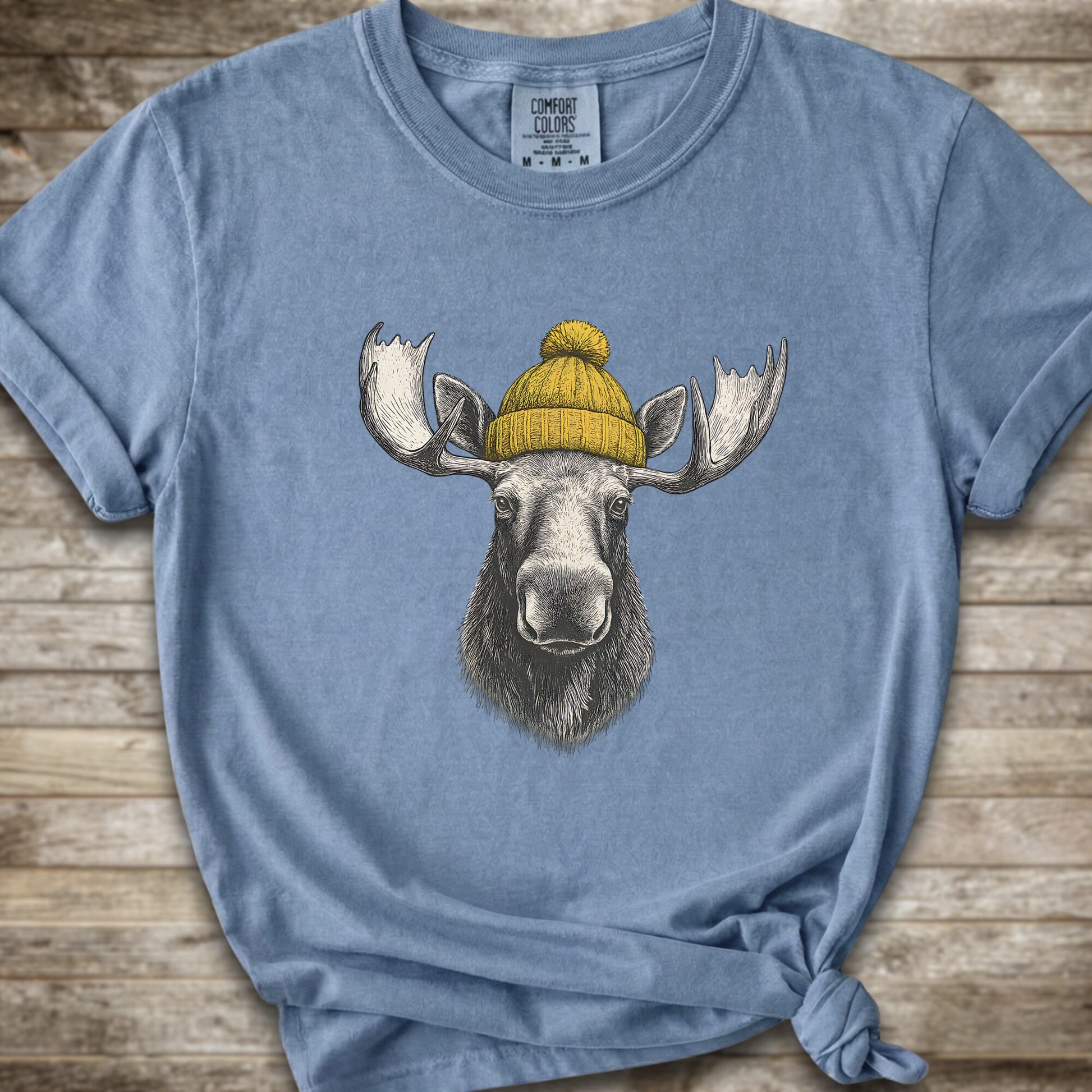 A light Blue short-sleeve t-shirt with a graphic of a moose wearing a yellow knit beanie, displayed on a rustic wooden background.